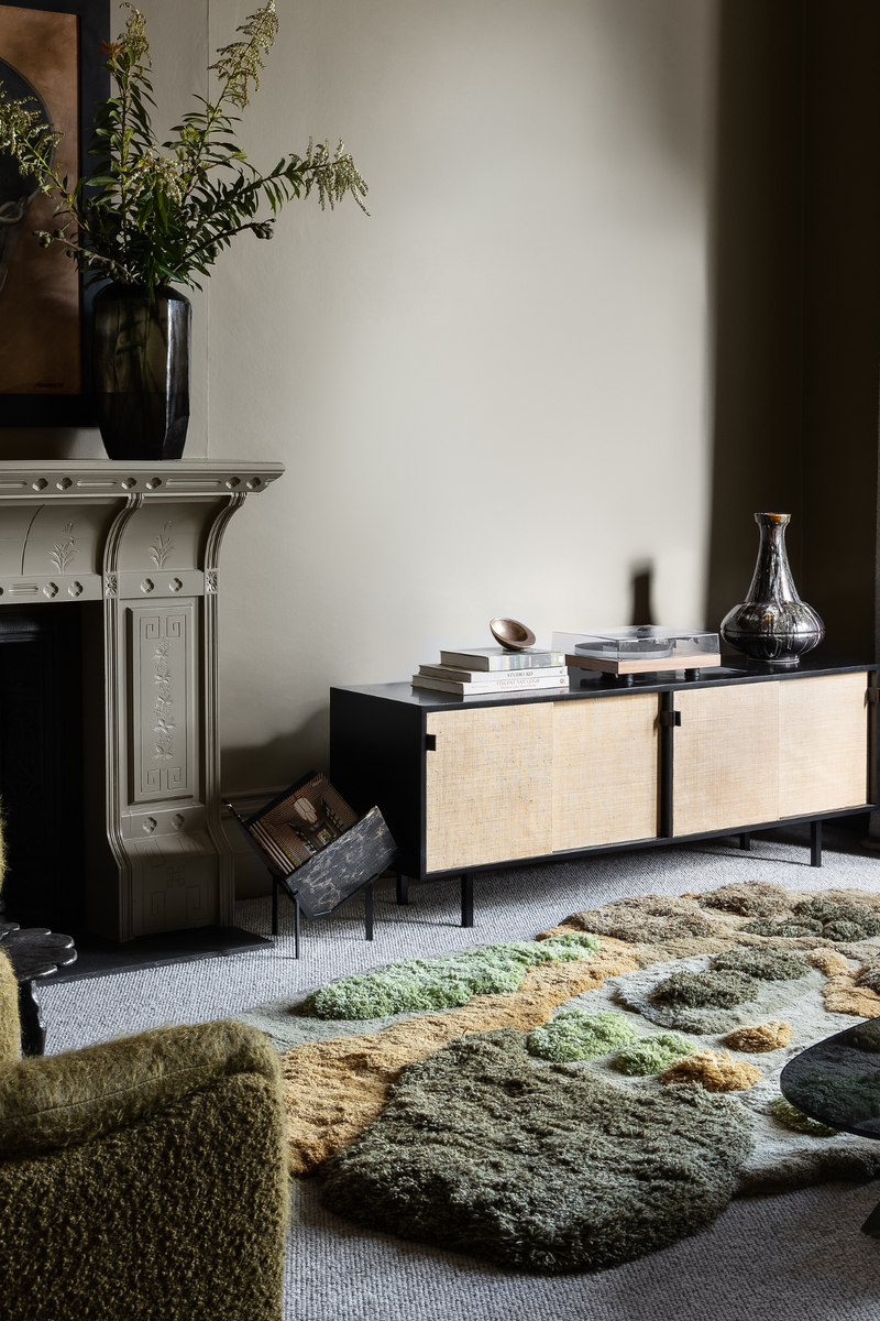 Cosy winter living room with textured green-toned rug, rattan sideboard, and a styled mantelpiece with leafy foliage.