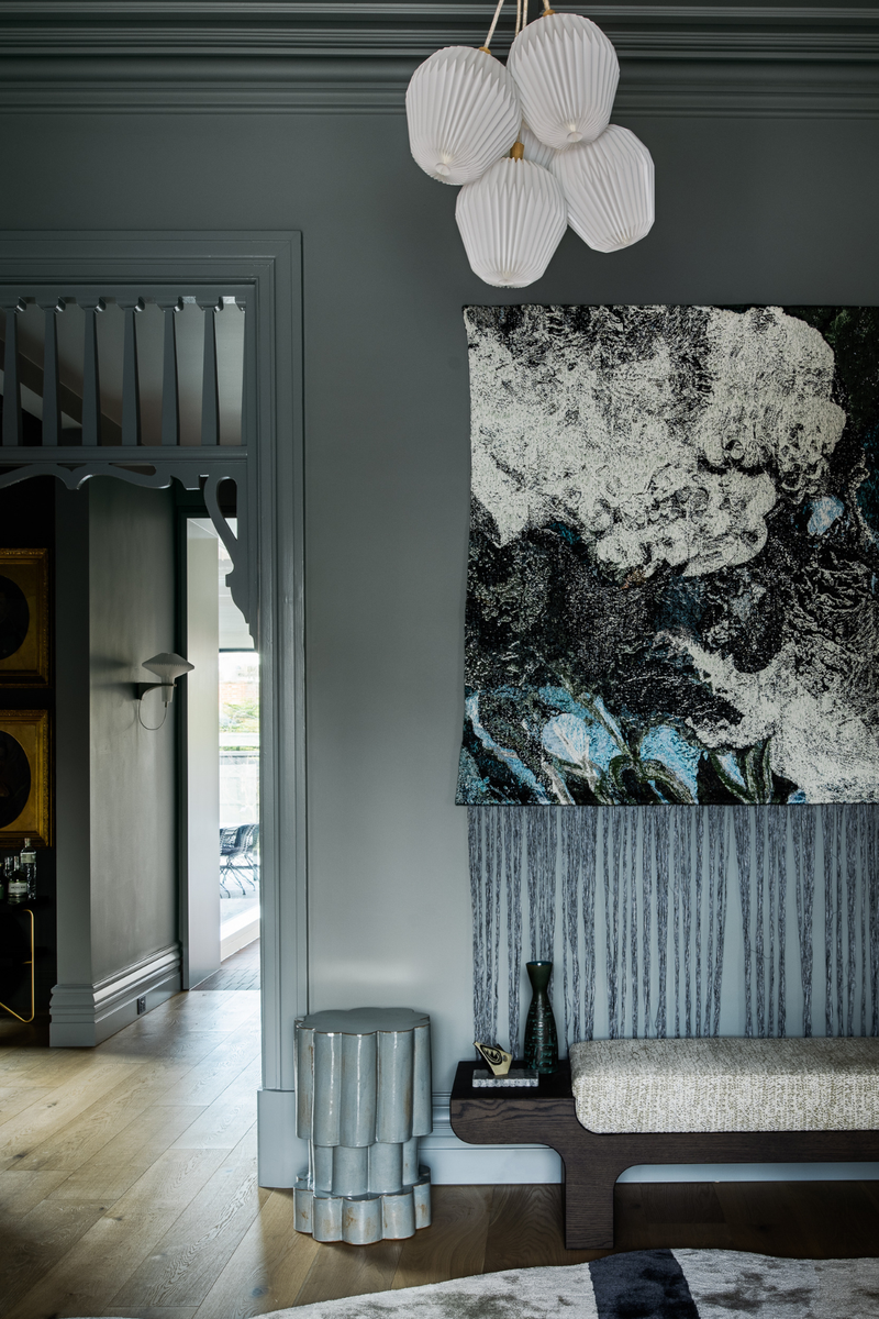 Moody interior with dark olive walls, ceramic pedestal side table, abstract wall hanging, and velvet-fringed artwork.