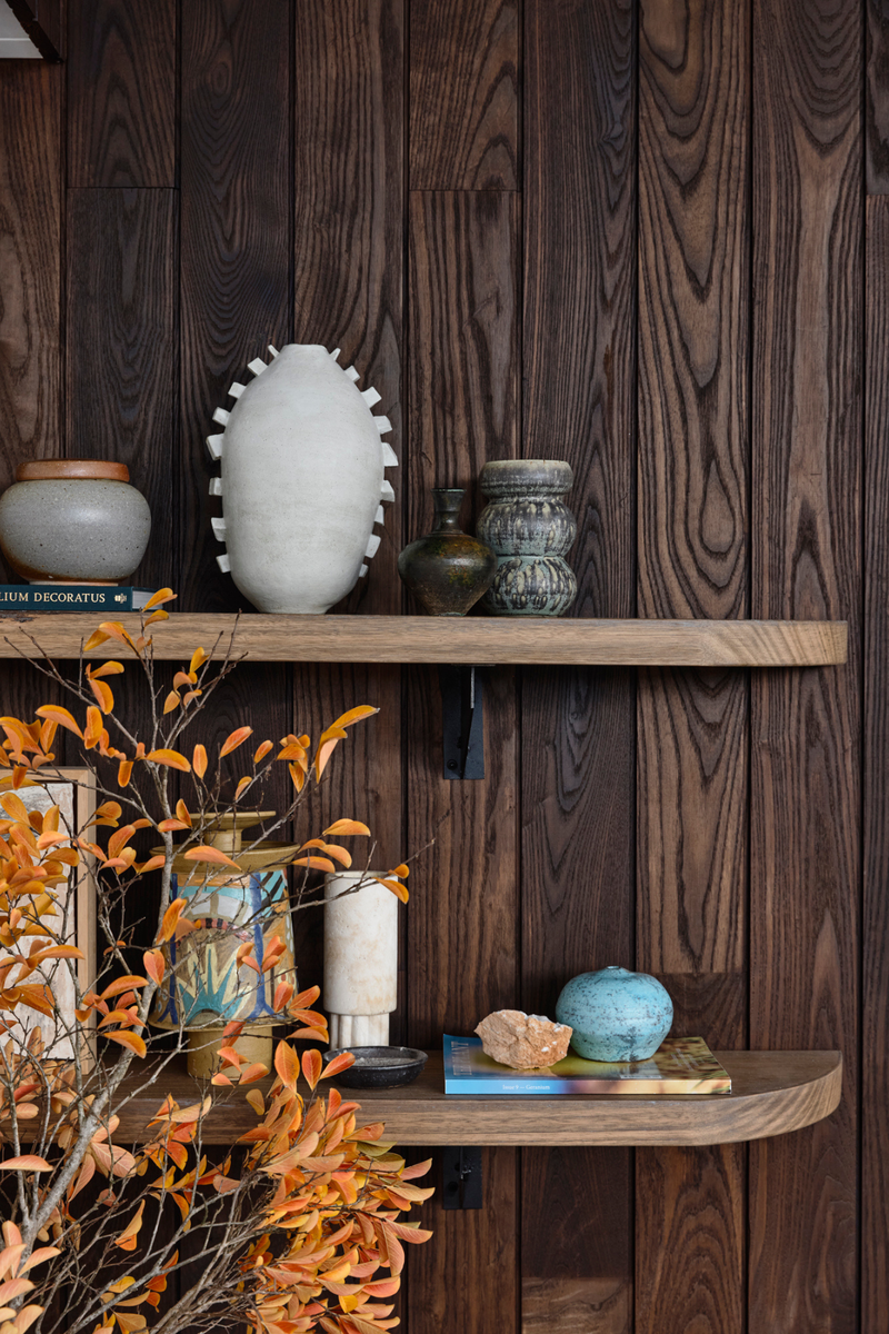 Curated timber shelf styling with ceramic vessels, books, and bare autumn branches against dark wood panelling.